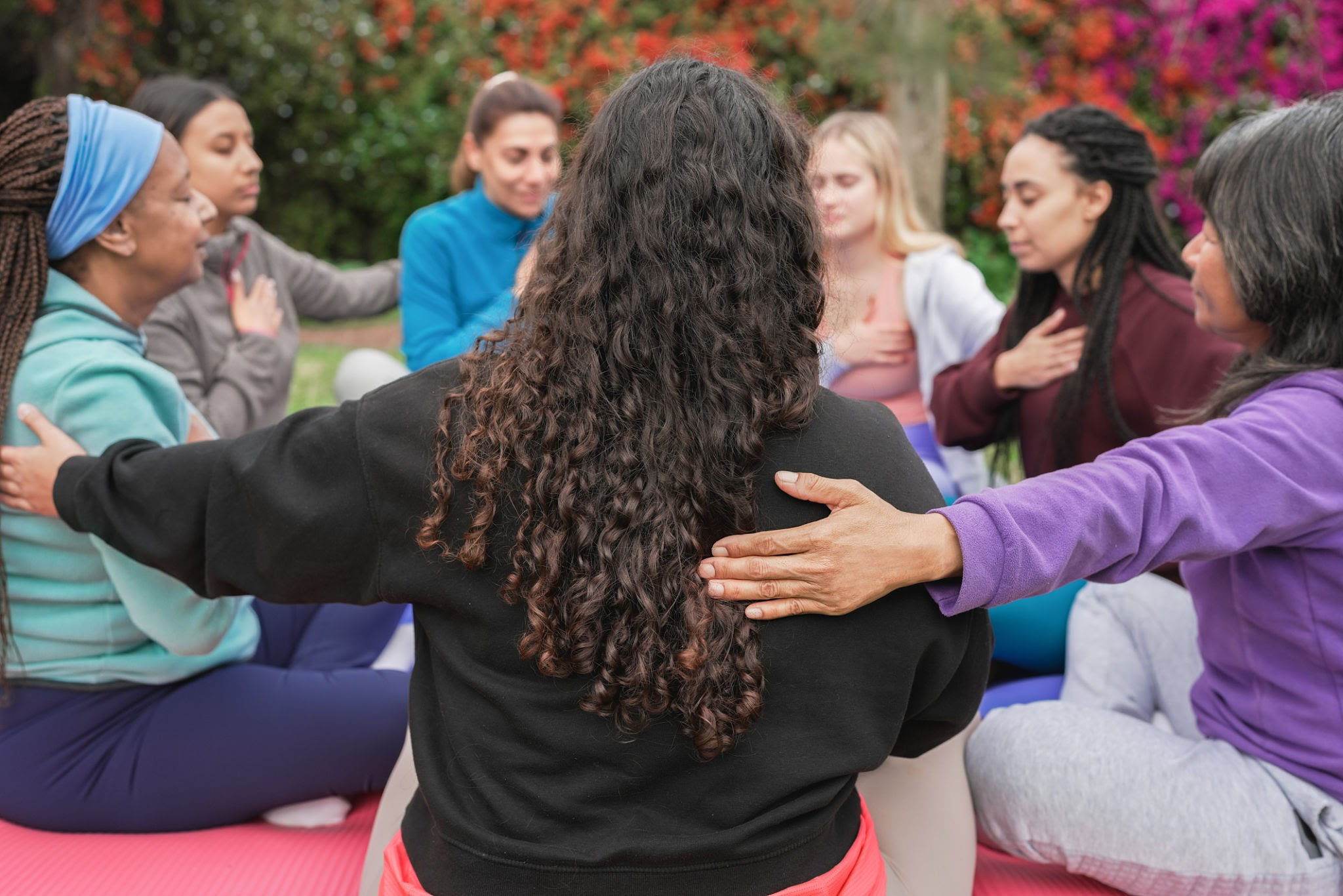 A group of women sitting in a supportive circle outdoors on yoga mats, with their eyes closed in meditation